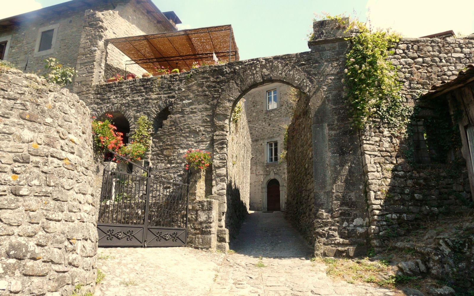 Arco de pedra entre construções antigas, com flores e vegetação