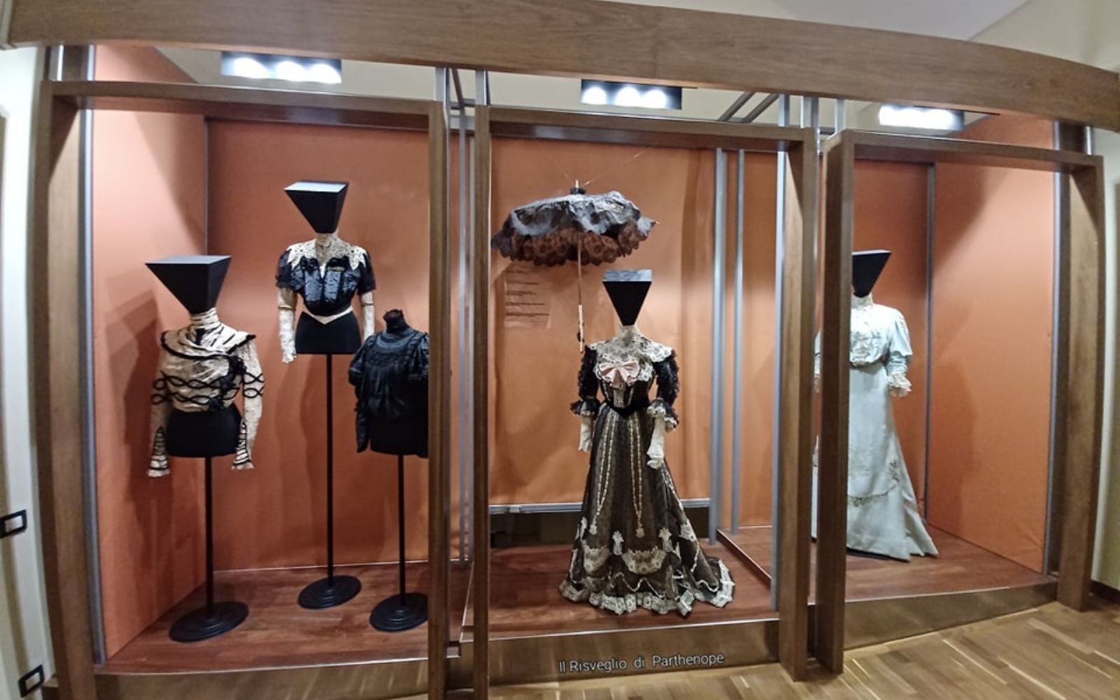 Vitrine com vestidos históricos, blusas e uma sombrinha no Museo della Moda di Napoli