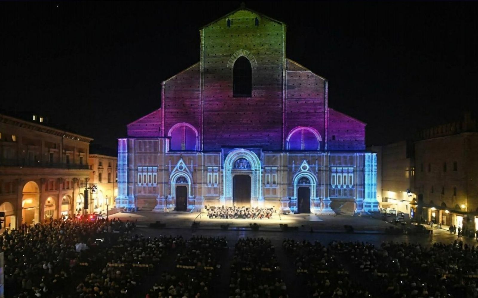 Fachada de San Petronio iluminada por projeções coloridas, com público na Piazza Maggiore à noite